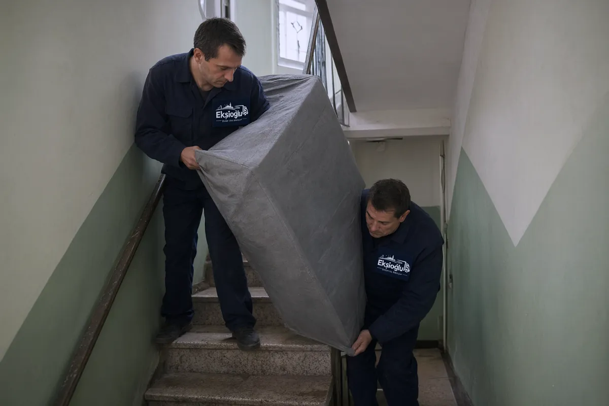Asansörsüz eski binada iki taşıyıcı battaniyeye sarılı gardırobu dar ve dönemeçli merdivenden indiriyor